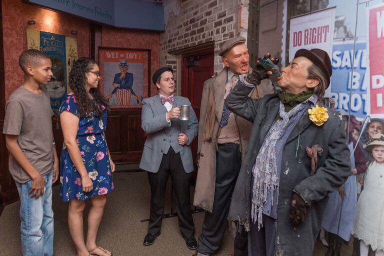 Wax likenesses of 2 drunkards next to a guy with a jacket and bowtie holding a mug who is standing next to two visitors to the American Prohibition Museum in Savannah, GA