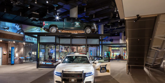 national law enforcement museum exhibit featuring a police car in the center with a door open and the words 'Indiana State Police' and in the background, glass case with a truck mounted on the top